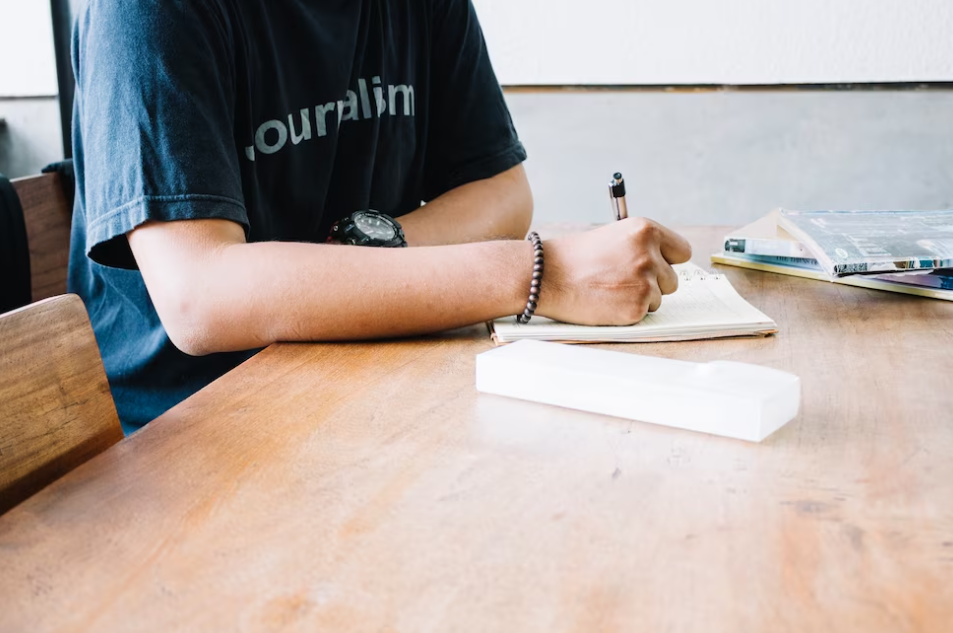 The person in a black t-shirt sits at the table and makes notes with a pen